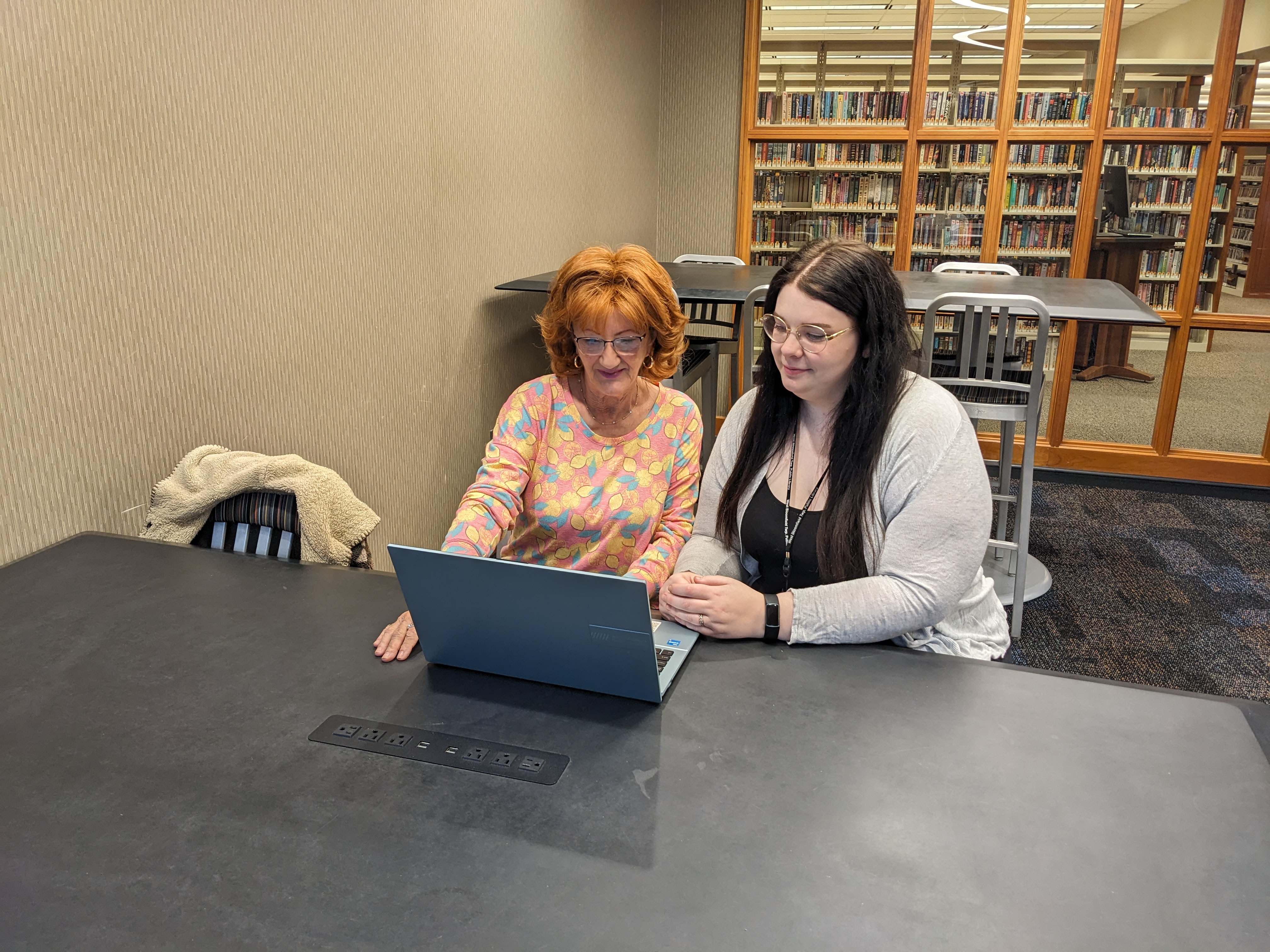 Two women look at a computer while seated at a table in the library