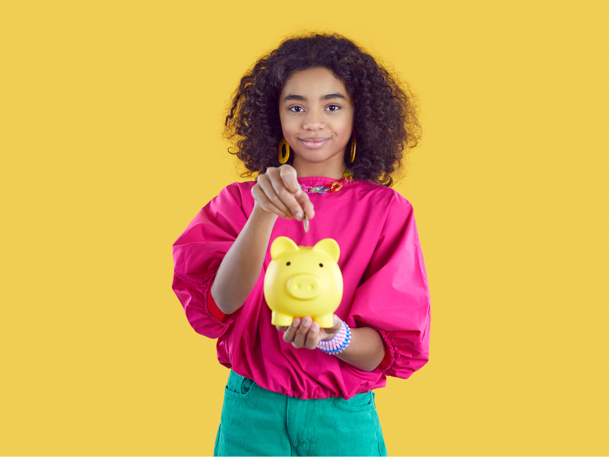 A tween aged person wearing a bright fuchsia shirt and turquoise pants is holding a yellow piggybank in front of them and placing a coin in it. The background is a mustard yello. 