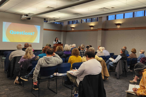 People sit in chairs in the Main Library meeting room. They face a speaker at a podium and a projector screen with the word "Questions?" on it.