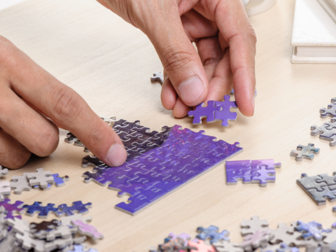 Two adult hands putting a mostly purple puzzle together on a table.