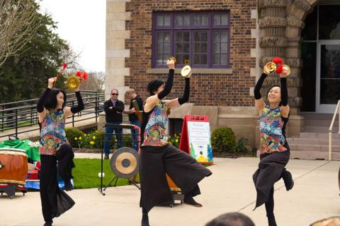 photo of three people dancing with traditional Japanese attire outside. In the background is a brick building with two men standing in front of it. 