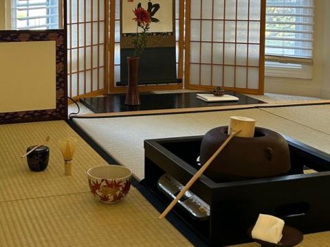 Photo of tatami mat with a box holding Japanese tea ceremony things inside, including a large pot and bamboo ladle. Also other tea ceremony bowls on the mat. The background has a screen and a plant in a vase in front of it.