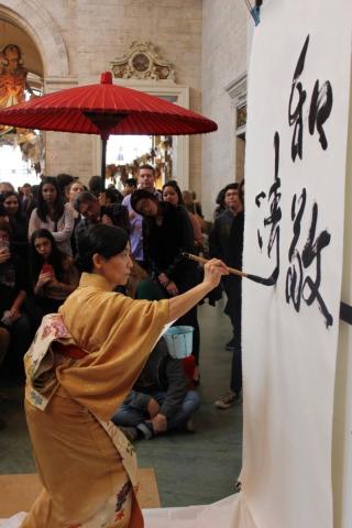 A person in a yellow kimono with a calligraphy brush is using it to write characters on a hanging white sheet of paper. There are people in the background watching.