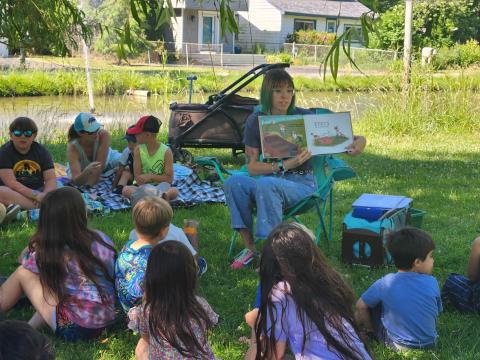 Ms. Mary reading a picture book under a willow tree as kids watch from blankets