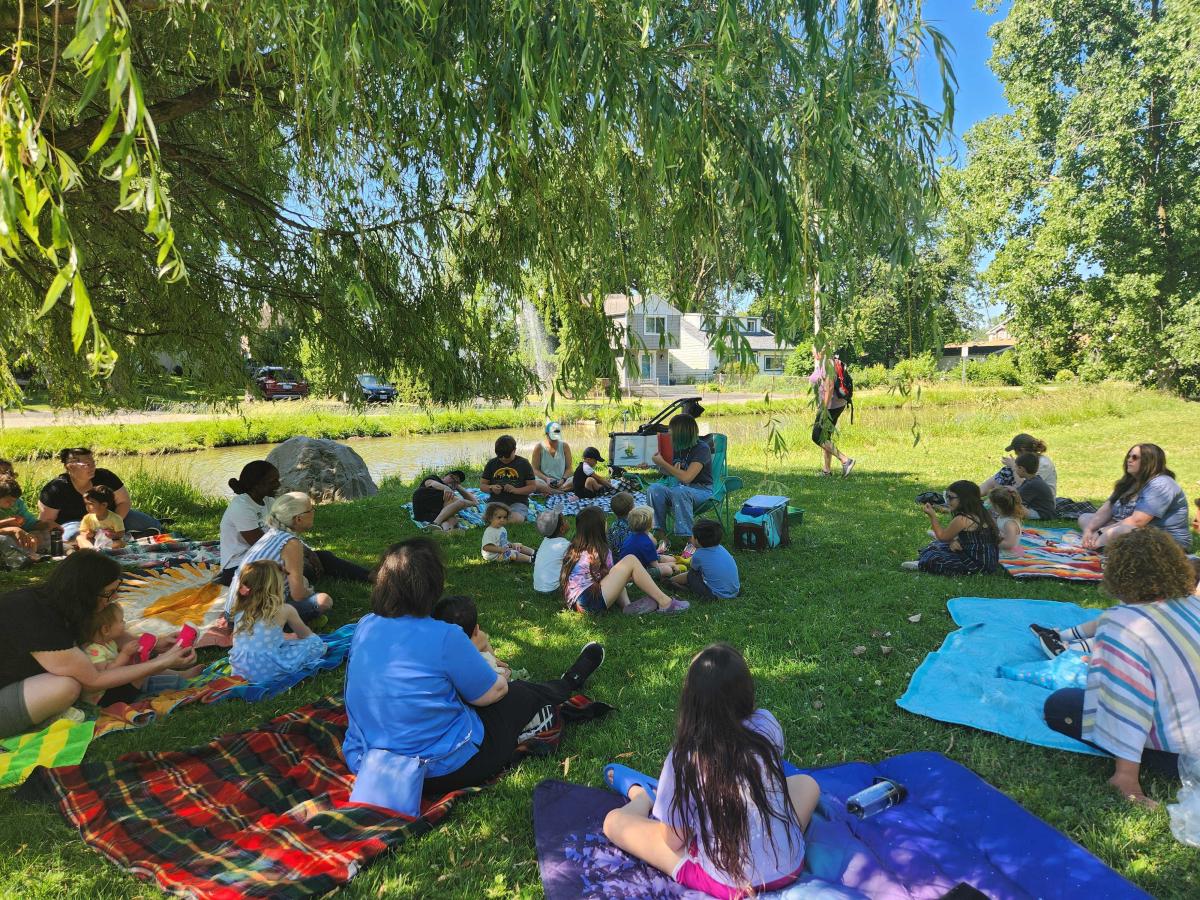 Ms. Mary reading a picture book under a willow tree as kids watch from blankets